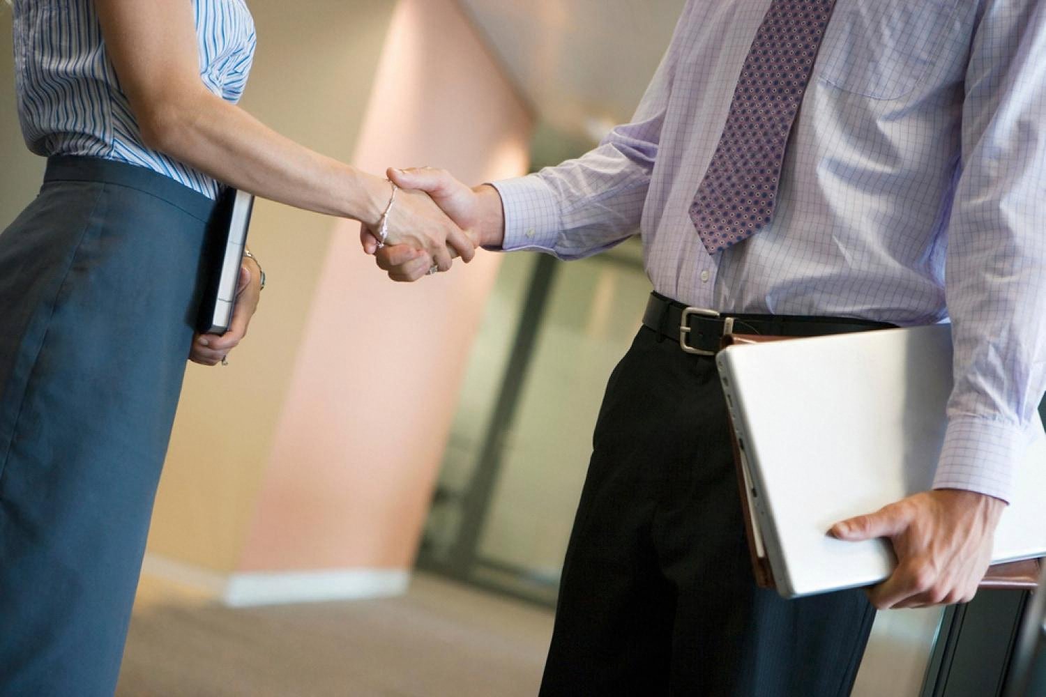 Two business professionals shaking hands in an office corridor after signing a compliance agreement